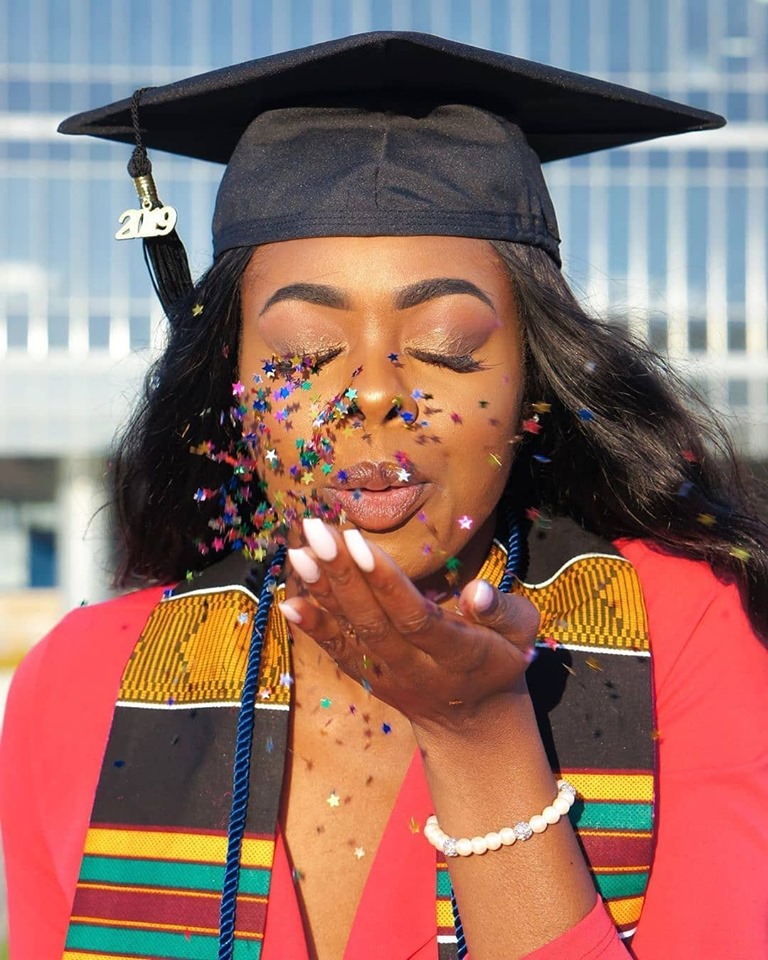 African American woman with long black hair is blowing sparkly confetti out of her right hand while wearing a black graduate cap with 2019 tassle, a red v-neck sweater, and kente cloth stole around her neck. 