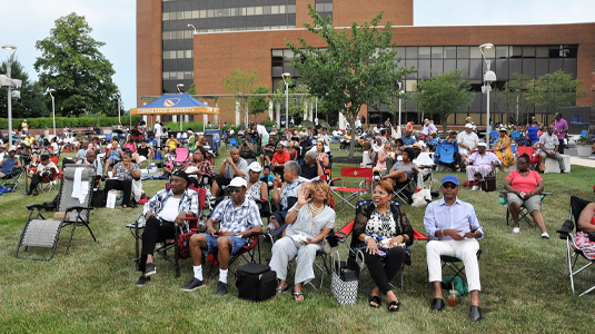 Large group of diverse people sitting in chairs on the grass in front of a Coppin campus building.