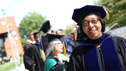 Brown skin woman wearing glasses and faculty regalia standing in the sunshine outside a campus facility.