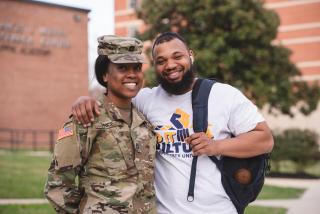 A Black female Army Cadet in uniform stands next to a Black male student with a short beard, who is wearing a white t-shirt with his backpack slung over his left shoulder.