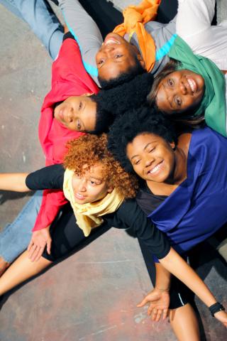 5 brown-skin young women each wearing a brightly colored shirt sitting closely grouped together with their arms outstretched and open