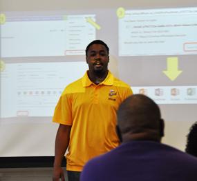 Brown skin man with short black hair wearing a yellow collared shirt stands in front of a projector screen.