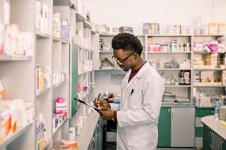 Young male student standing in interior of pharmacy and making notes on clipboard.