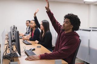 Students sit at workstations in the computer lab. Two students are raising their hands to ask question.