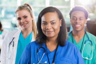 Young African American female nurse stands in front of Caucasian physician and African American male nurse. They are wearing scrubs and lab coats and are looking at the camera.
