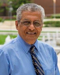 Brown skin man with short gray hair and wire-rim glasses wears a blue button down shirt with striped tie while standing in front of a Coppin State University campus building.