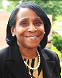 Brown skin woman with shoulder-length black hair part on the right side, wearing a black blazer, string of pearls and earrings, with black and yellow striped blouse
