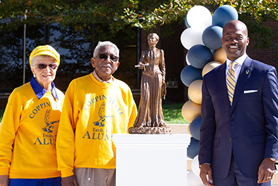 James "Winky" and Florine "Peaches" Camphor wear yellow Coppin alumni sweatshirts and sunglasses while standing next to President Anthony Jenkins and a miniature bronze statue of Fanny Jackson Coppin