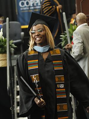 Brown skin woman wearing glasses, black graduation robe, cap, and kente cloth stole walks from the graduation stage with her degree in her right hand