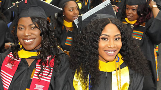 2 smiling students wearing black graduation caps & gowns under colorful stoles.