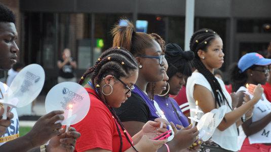 Group of brown skin people with diverse hairstyles holding candles outside a campus building.