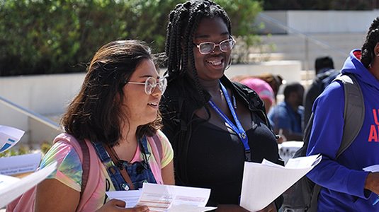 2 students both wearing glasses and holding papers while standing outside a campus building in the sunshine.