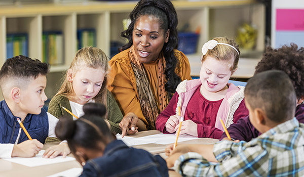 A mature African-American woman teaching a group of six multi-ethnic elementary school children. They are sitting around a table in the classroom. The students are writing as the teacher talks. The girl in the red shirt has down syndrome.