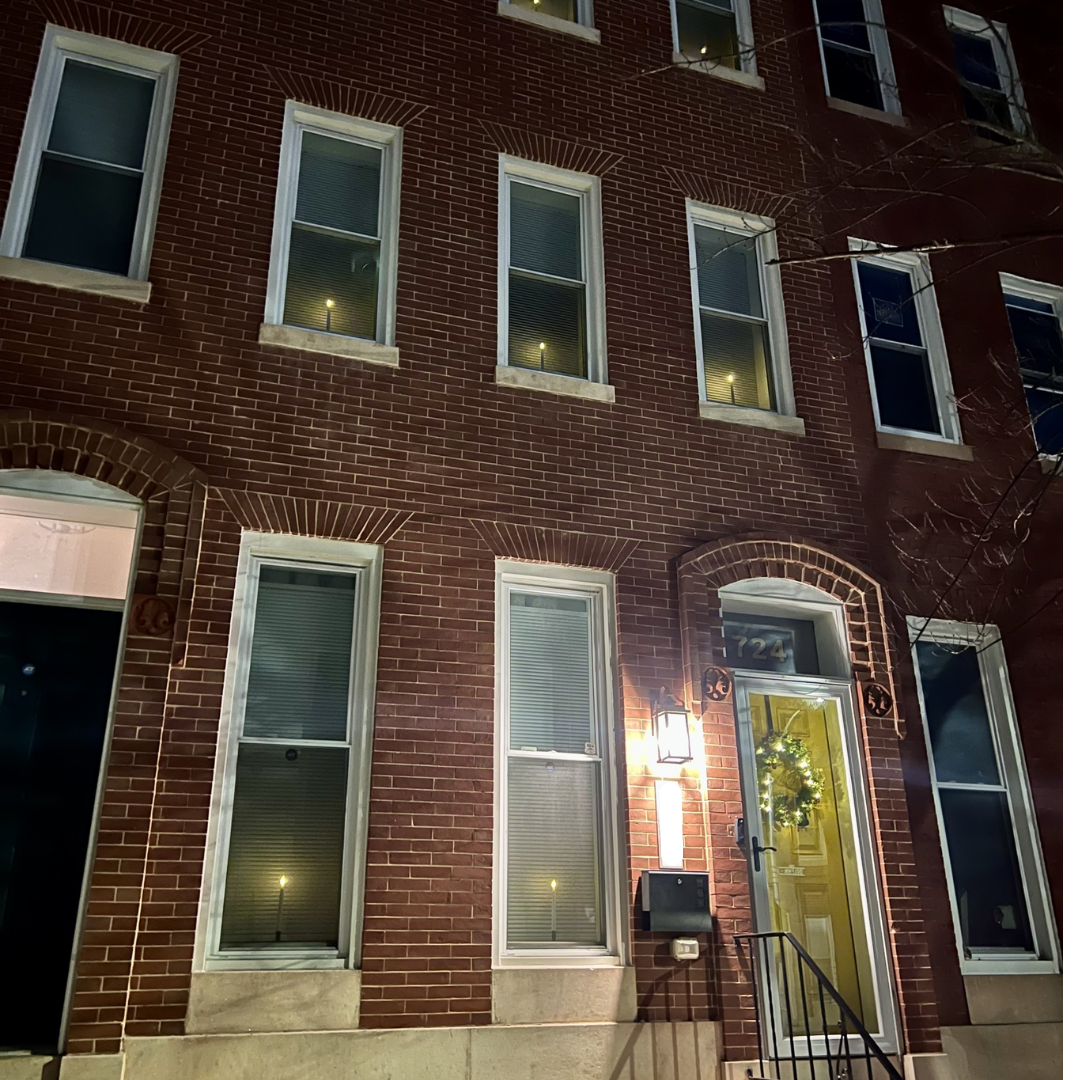 Classic Baltimore brick rowhouse photographed at night, featuring white-trimmed windows across three floors, a yellow front door with a decorative wreath, and a warm porch light illuminating the entrance.
