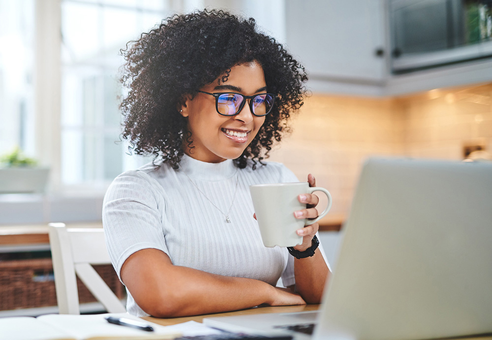 photo of a woman with curly hair and glasses, smiling while looking at a laptop and holding a mug