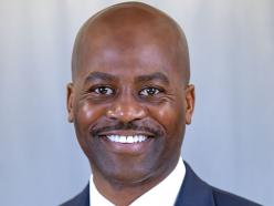 Headshot of CSU President Anthony Jenkins wearing a white collared shirt, bright yellow tie, navy blue suit jacket with CSU lapel pin in front of green shrubs and low brick wall on Coppin's campus