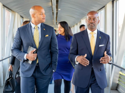 Four professionals walking together in a brightly lit corridor, two men in suits and two women, one smiling and wearing a blue dress_.png