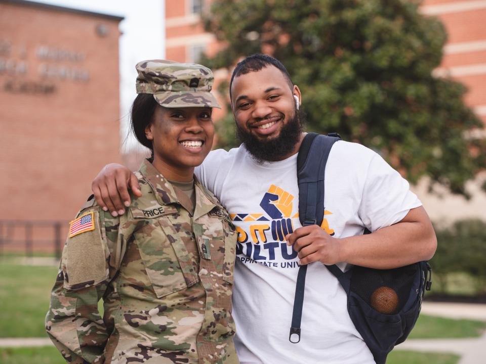 A Black female Army Cadet in uniform stands next to a Black male student with a short beard, who is wearing a white t-shirt with his backpack slung over his left shoulder.