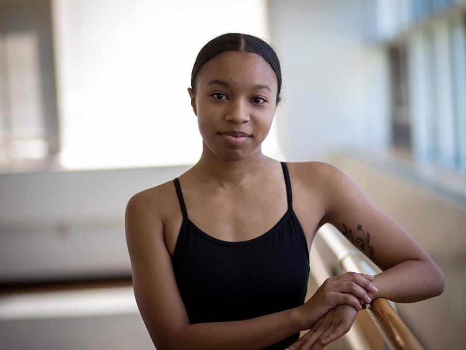 A dance student in a black leotard rests her arm on the barre in the dance studio
