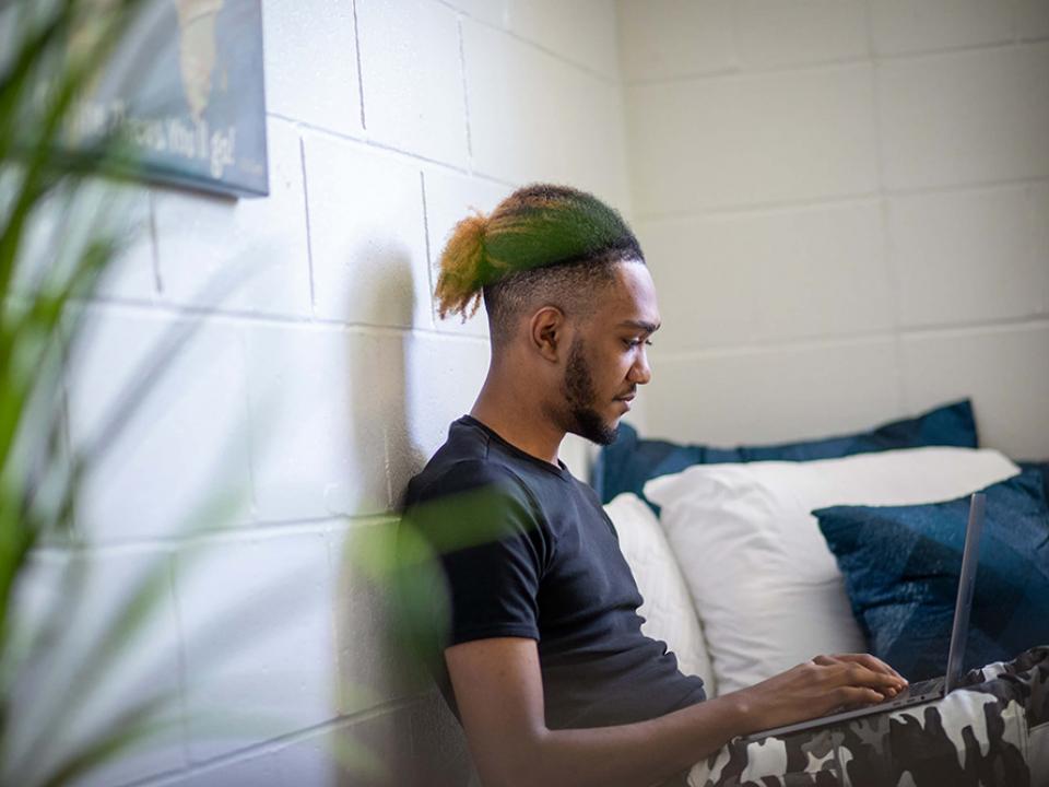 Male student sits on his dorm room bed working on a laptop