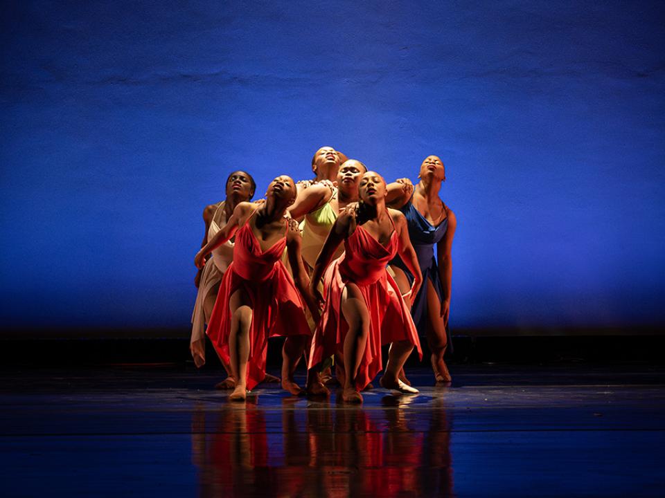 Ballet dance ensemble in flowing dresses performing a unified pose under blue stage lighting.