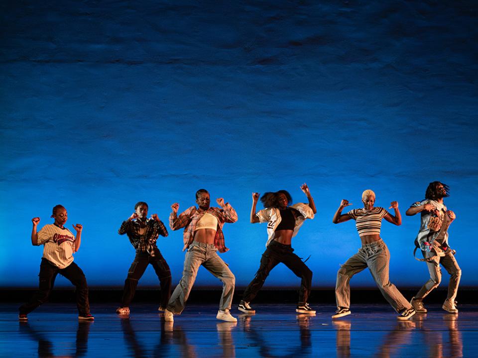 Street dance group in casual attire performing synchronized movements on a stage with a blue backdrop.