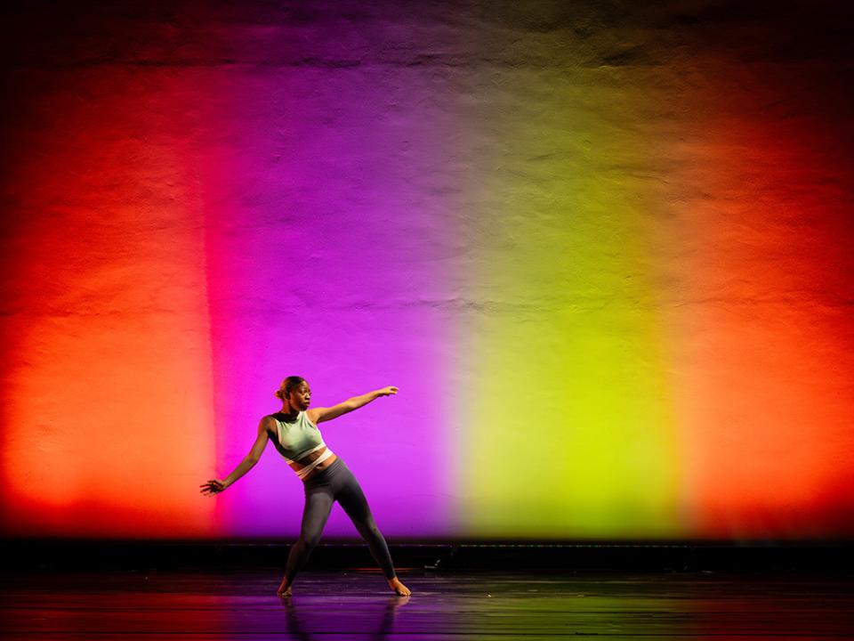 Solo dancer in activewear extending her arm on stage with vibrant red, purple, and yellow lighting.