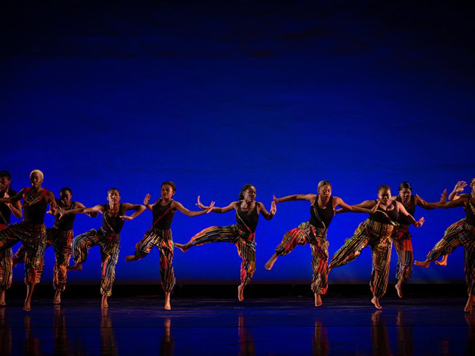 Group of dancers in African-inspired costumes mid-leap during a high-energy performance on a blue-lit stage.