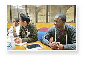 Three male students sitting on one side of a wooden table intently listening to a non-visible speaker