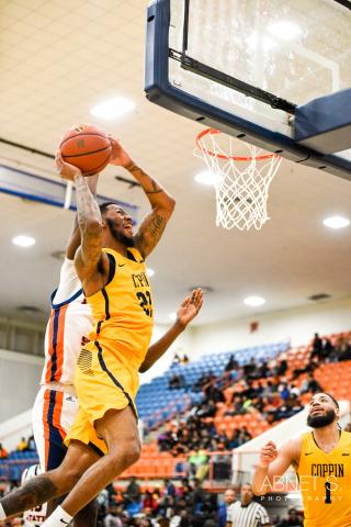 Brown skin man in yellow and black basketball jersey and shorts has basketball in both hands preparing to dunk in the net while defending against another player behind him in a white, orange in blue jersey.