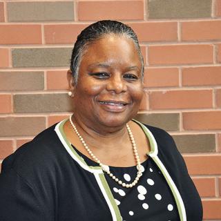 Brown skin woman with short gray and black hair wearing pearls with a black sweater trimmed in green and white with a black and white polka dot blouse while standing in front of a red-ish brick wall