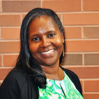 Brown skin woman with long black and silver hair smiles and is wearing earrings and a green and white patterned blouse with a black sweater while smiling and standing in front of a red-ish brick wall
