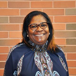 Brown skin woman with shoulder-length dark hair wearing glasses, stud earrings and a high neck blue patterned blouse while standing in front a red-ish brick wall