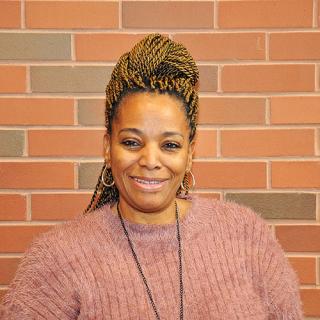 Brown skin woman with hair in long light brown twists wearing hoop earrings, black necklace, and pink ribbed sweater smiling in front of a red-ish brick wall.