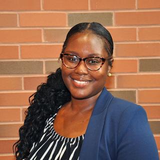 Brown skin woman with long wavy black hair wearing a navy blue blazer with a white and black striped blouse while standing in front of a red-ish brick wall