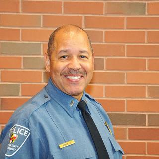 Brown skin man wearing a blue police officer's uniform smiling and standing in front of a red-ish brick wall