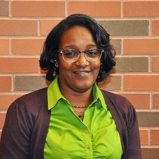 Brown skin woman with short black curly hair wears glasses with a necklace, neon green button-up blouse and maroon sweater stands in front of a red-ish brick wall