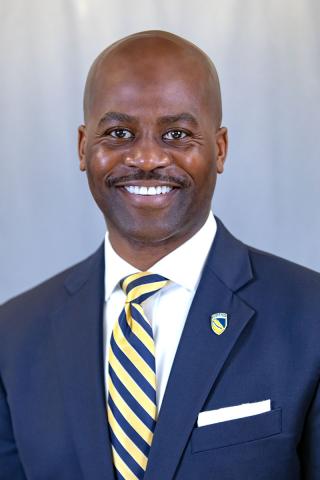 Headshot of CSU President Anthony Jenkins wearing a white collared shirt, bright yellow tie, navy blue suit jacket with CSU lapel pin in front of green shrubs and low brick wall on Coppin's campus