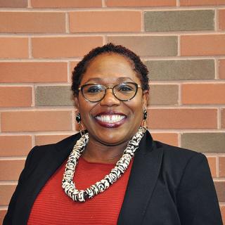 Brown skin woman with short curly dark hair smiles while wearing stud earrings, a white and black necklace, red blouse and black blazer while standing in front of a red-ish brick wall
