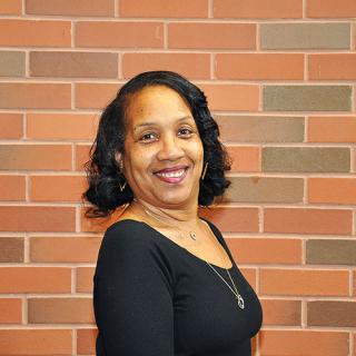 Brown skin woman wearing a black scoop neck top with long silver necklace standing with right shoulder while smiling and standing in front of a red-ish brick wall