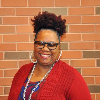 Smiling brown skin woman wearing glasses, hoop earrings, silver necklace, red sweater with red and blue patterned blouse stands in front of a red-ish brick wall