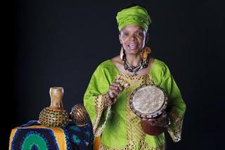 A senior African American storyteller wearing colorful African clothing and jewelry. She is holding an African drum and is in the midst of a story.
