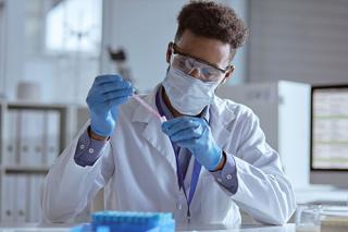 A male in lab coat and goggles drops liquid into a test tube