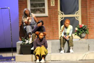 Four Black women sit on the front steps of a brick house in a stage play, each looking tired, with flowers and a screen door behind them.