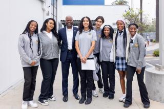 A group of eight people, including seven students and one adult male, posing for a photo outdoors in front of a white building. The adult, positioned third from the left, wears a navy blue suit and a white dress shirt. The students are dressed in school uniforms consisting of grey polo shirts, sweaters, or cardigans with a crest, paired with dark trousers or a blue plaid skirt. 