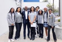 A group of eight people, including seven students and one adult male, posing for a photo outdoors in front of a white building. The adult, positioned third from the left, wears a navy blue suit and a white dress shirt. The students are dressed in school uniforms consisting of grey polo shirts, sweaters, or cardigans with a crest, paired with dark trousers or a blue plaid skirt. 